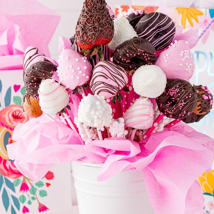 A beautiful bouquet of chocolate covered strawberries in a white pail with pink tissue paper and gift bags in the background