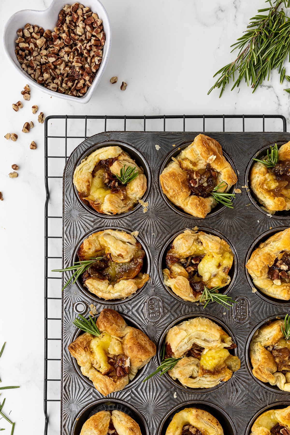 antique muffin pan filled with baked brie bites on black cooling rack sitting on a white marble countertop with rosemary and a heart shaped dish full of walnuts in the back