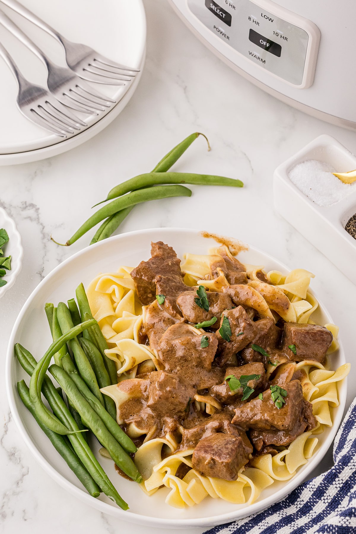 a dinner plate with egg noodles, beef tips in gravy, and green beans on a white marble counter, with salt and pepper wells, a crock pot, and plates with three forks in the background