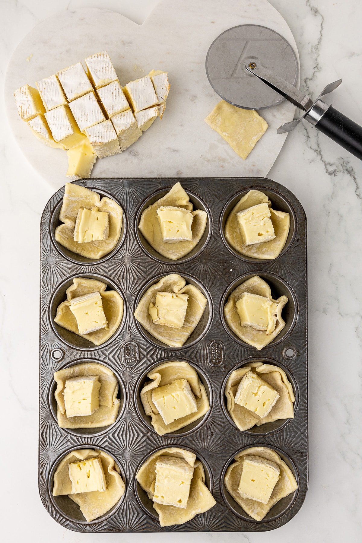 antique muffin pan on a white marble countertop with puff pastry squares and cubes of brie in each cup. Pizza cutter in the background