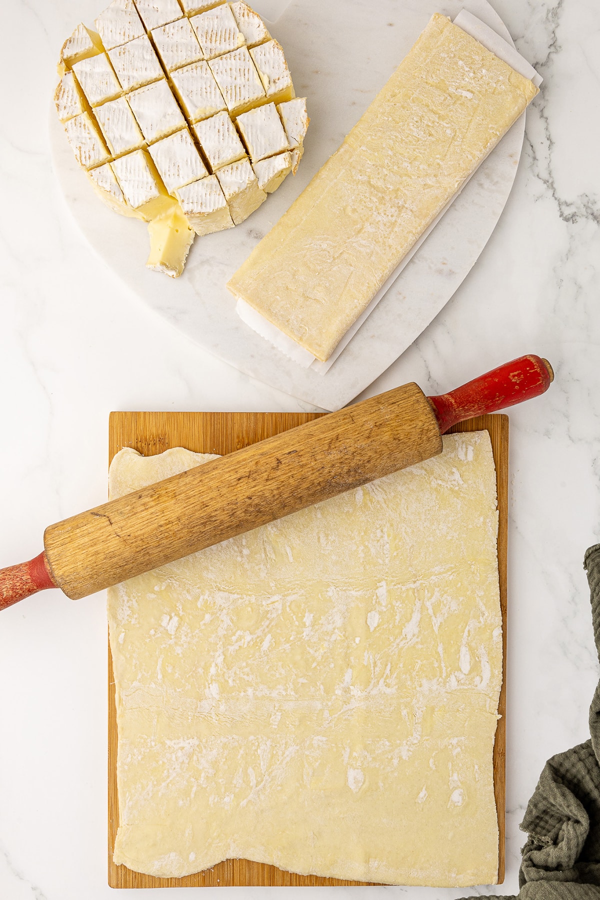 puff pastry being rolled out with a rolling pin on a wood cutting board, with cubed brie and another puff pastry sheet in the background