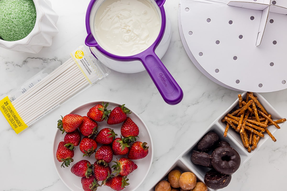 A plate of fresh strawberries on a white countertop by melted white chocolate, sucker sticks and other treats for dipping