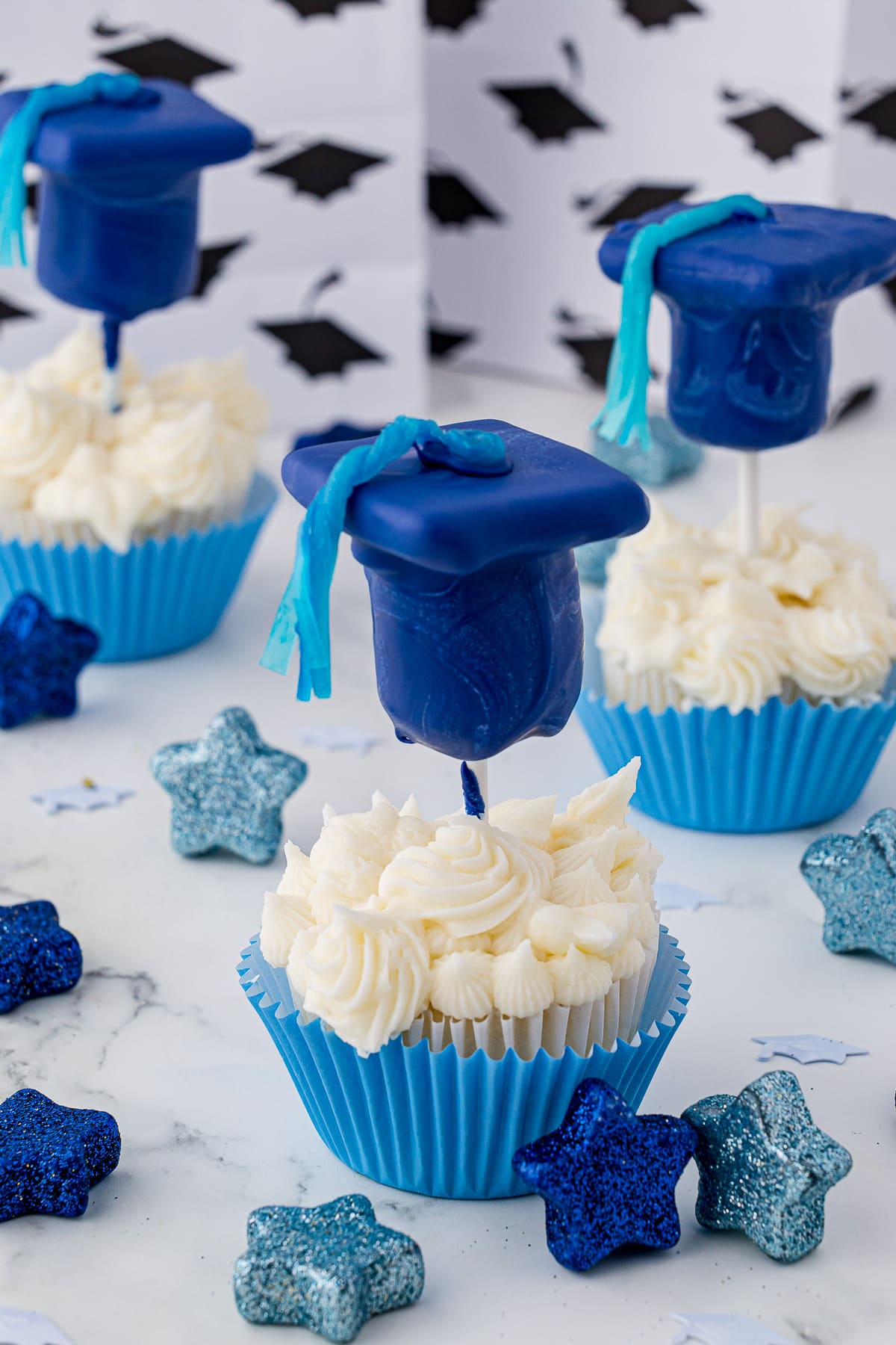 A white cupcake with white frosting in a blue cupcake wrapper topped with a blue chocolate graduation cap and a blue tassel made out of fruit rollups