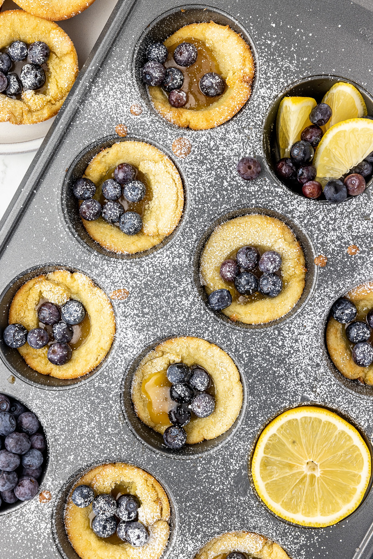 dutch babies in a cupcake tin with lemon curd, blueberries and lemon slices sprinkled with powdered sugar