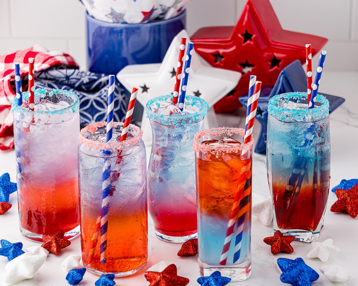5 glasses of red, white and blue layered drinks in tall thin glasses, and surrounded by red, white and blue decor including one red, one white and one blue star standing behind the drinks, and small red white and blue stars surrounding the drinks, along with red and white linen and blue and white linen. Each drink has a blue and white straw and a red and white straw in it