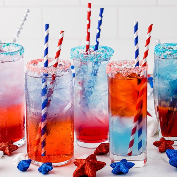 5 glasses full of red, white and blue layered drinks, with two straws in each glass, each glass rimmed with pop rock like candy; and, red, white and blue star decor surrounding the glasses.