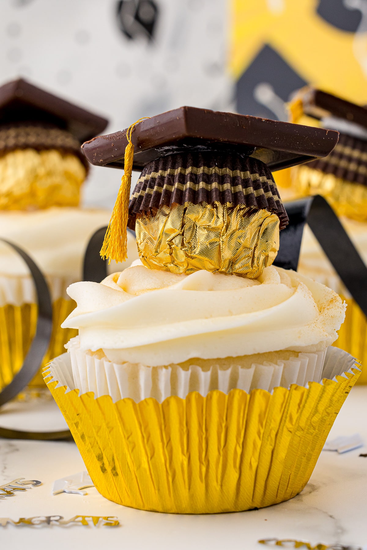 Close up of a white cupcake with white frosting in a gold cupcake wrapper and topped with a chocolate graduation cap and tiny told tassel.