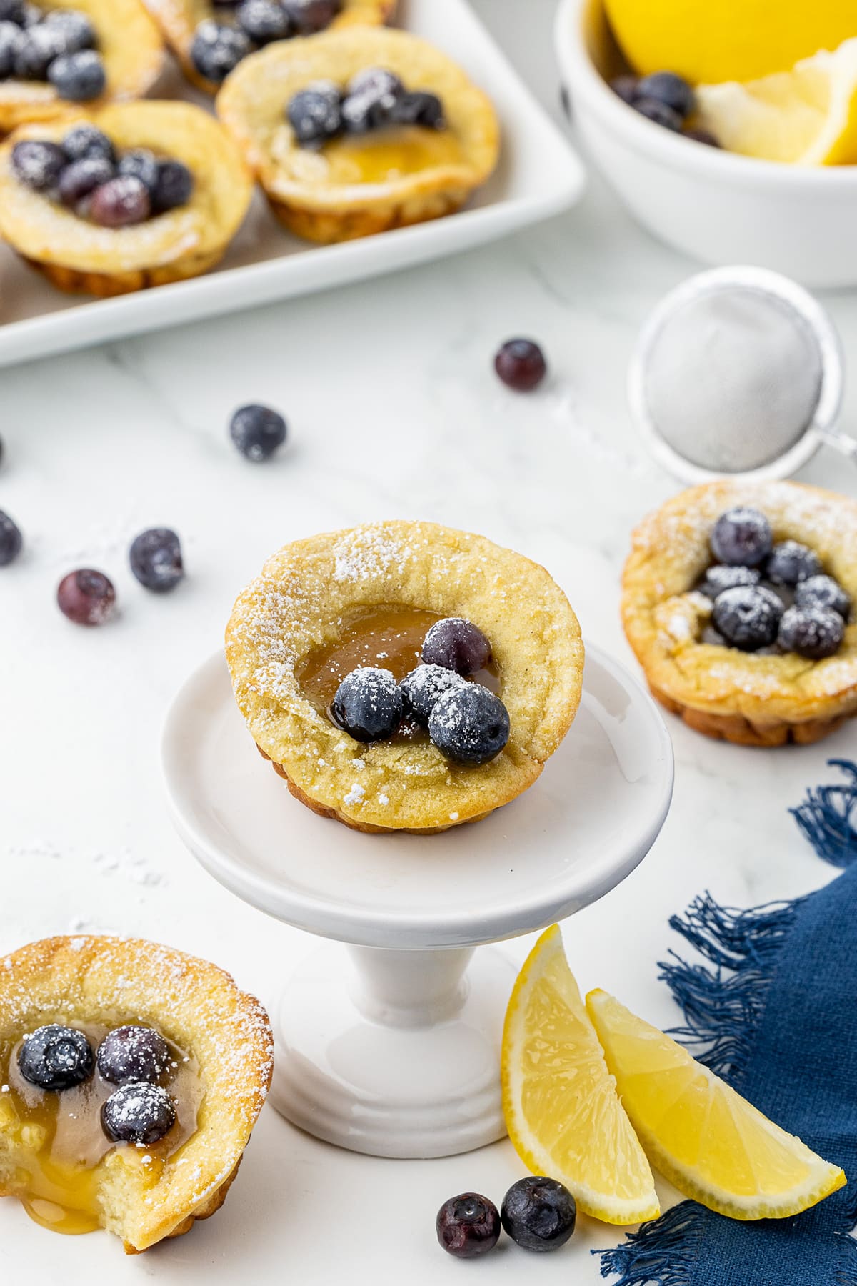 mini dutch baby on a white pedestal surrounded by other pancakes, lemon slices, and a blue napkin