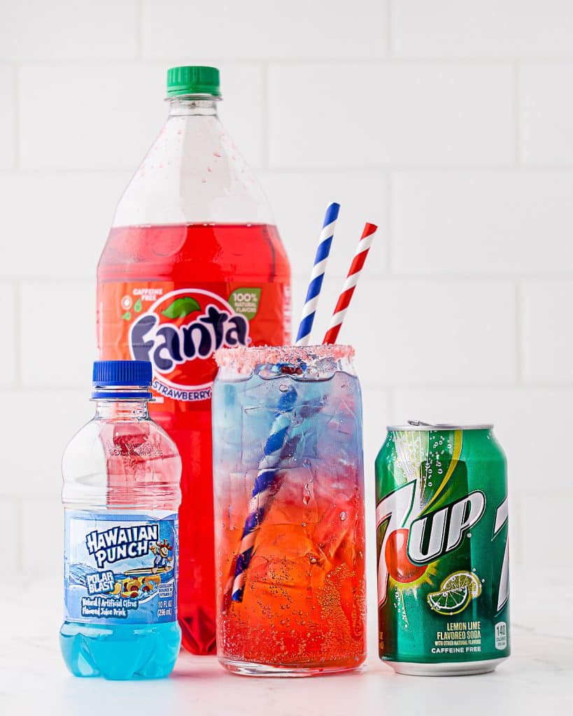 Red white and blue drink in round tall glass with blue Hawaiian punch, 7Up, and Strawberry Fanta. Glass has 2 straws, one with blue and white spirals and the other with red and white spirals