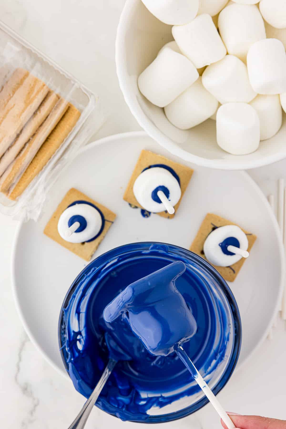 A white bowl of large marshmallows and a tray of graham crackers to be dipped into a bowl of melted blue chocolate