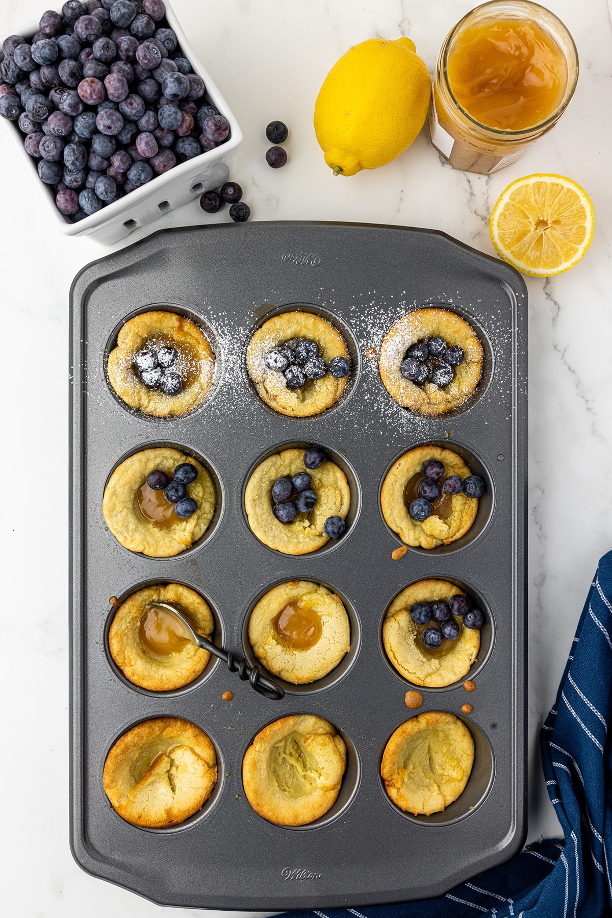 partially assembled mini dutch babies in a wilton cupcake tin with a berry basket filled with blueberries, lemon curd in a jar, and two lemons, on a white marble countertop