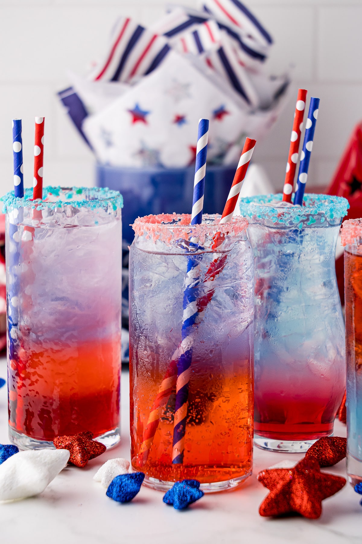 3 glasses of red white and blue layered drink with 2 straws each, rimmed with pop rock like candy, and decorated with red white and blue star decor, and red, white and blue napkins