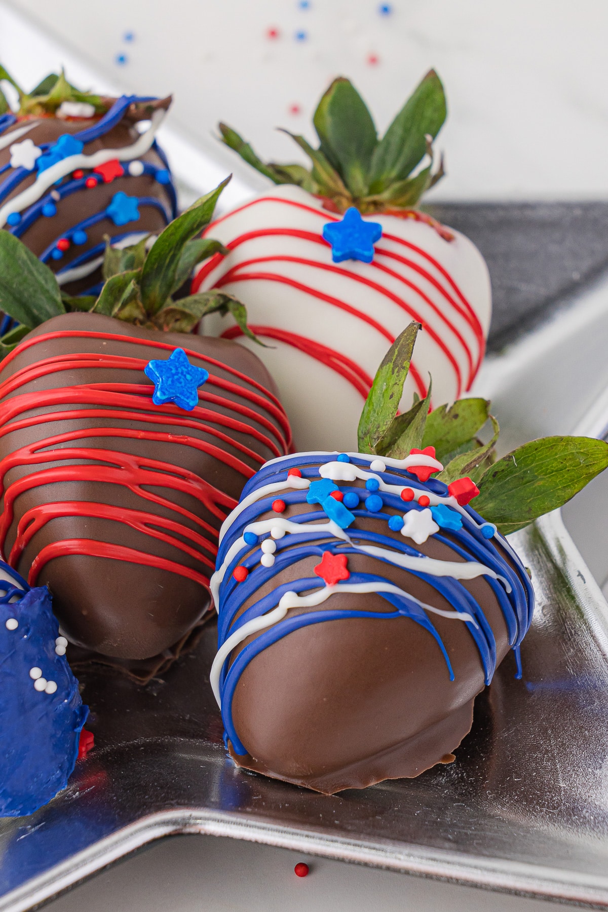 Close up of 5 chocolate covered strawberries plated on a silver star dish, and with red, white and blue edible striping, stars and sprinkles