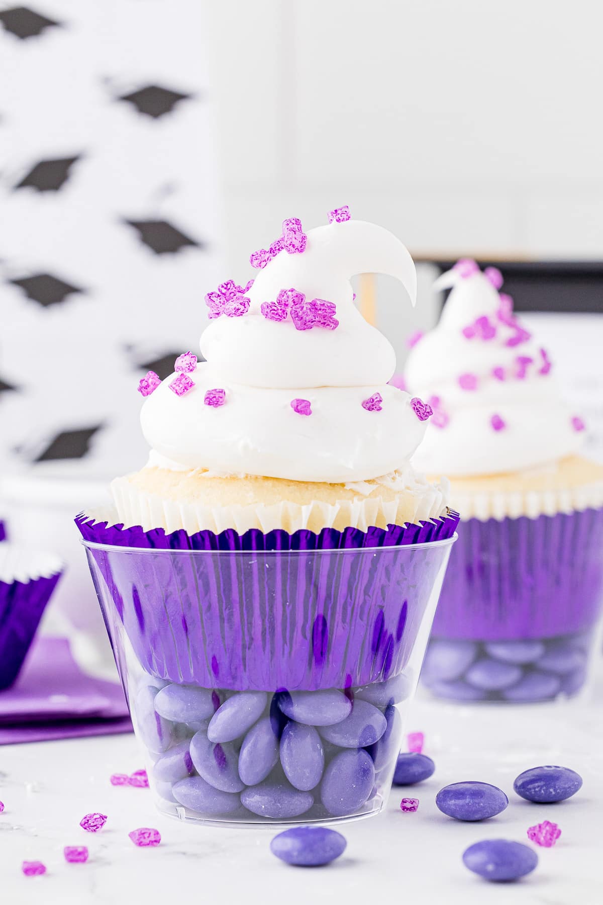 A cupcake in a purple foil wrapper sitting in a clear plastic cup on top of purple M&Ms topped with white frosting and sugar sprinkles