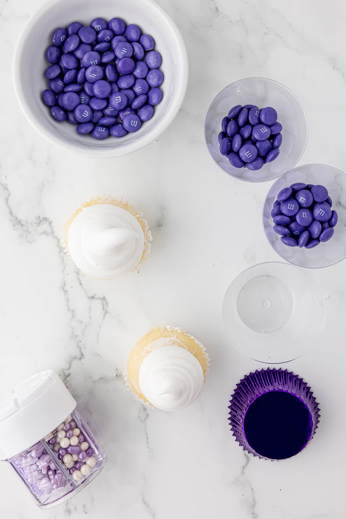 Bowls of purple M&Ms sitting on a marble countertop with undecorated cupcakes and a container of sprinkles