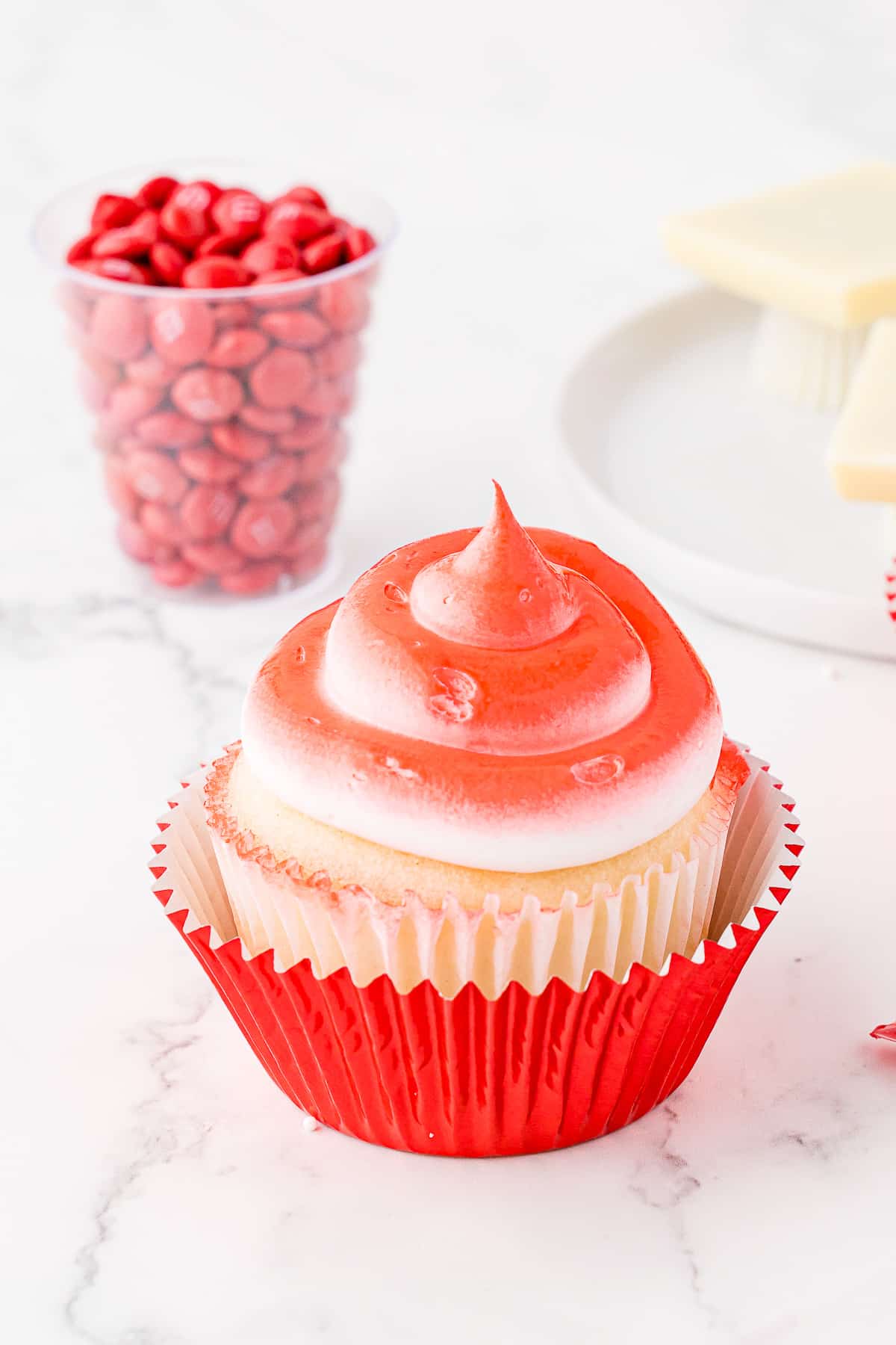A cupcake with sprayed red frosting in a red foil cupcake wrapper with a clear glass of red M&Ms behind it