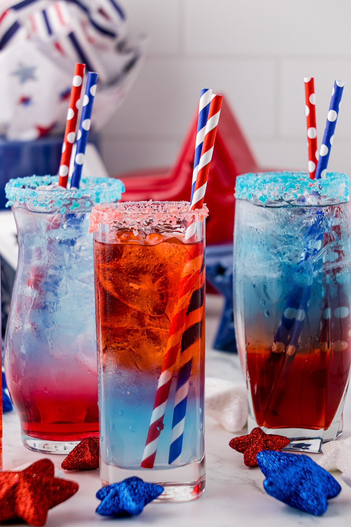 Three tall thin glasses with a red, white and blue layered drink; two with red at the bottom and one with blue at the bottom. All with two straws and each with pop rock like candy around the entire rim of glass
