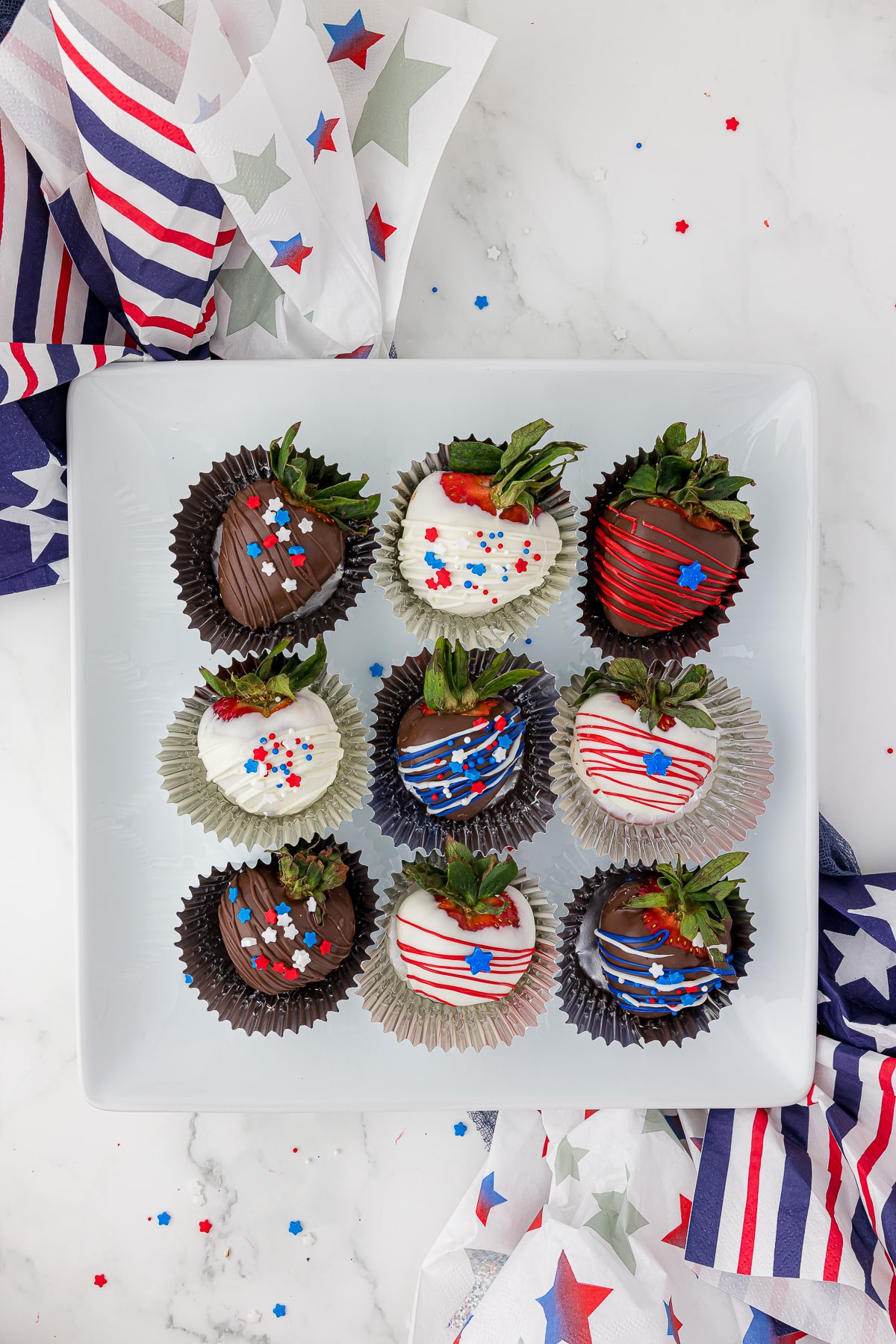 9 chocolate covered strawberries decorated with red, white and blue candies and stripes, plated on a square white dish, surrounded by red, white and blue napkins