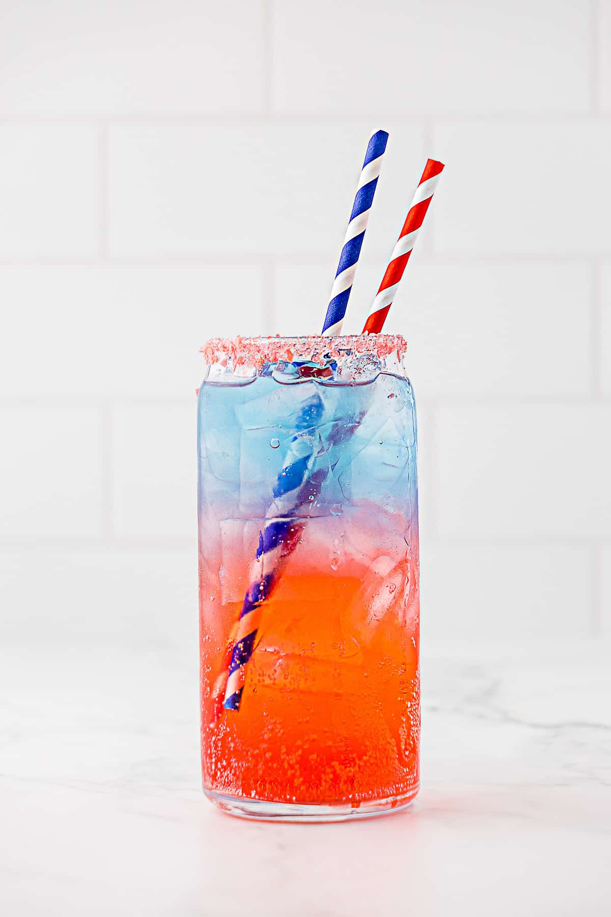 Layered red white and blue drink in tall thin glass with two straws and pop rock candy on rim of glass