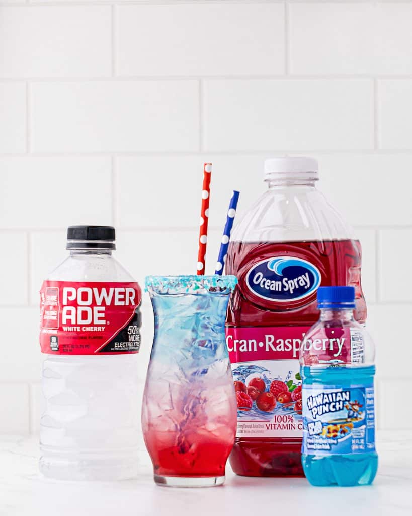 Layered red, white, and blue drink in an hour shaped glass with powerade, cran-raspberry, and Hawaiian punch bottles in the background.