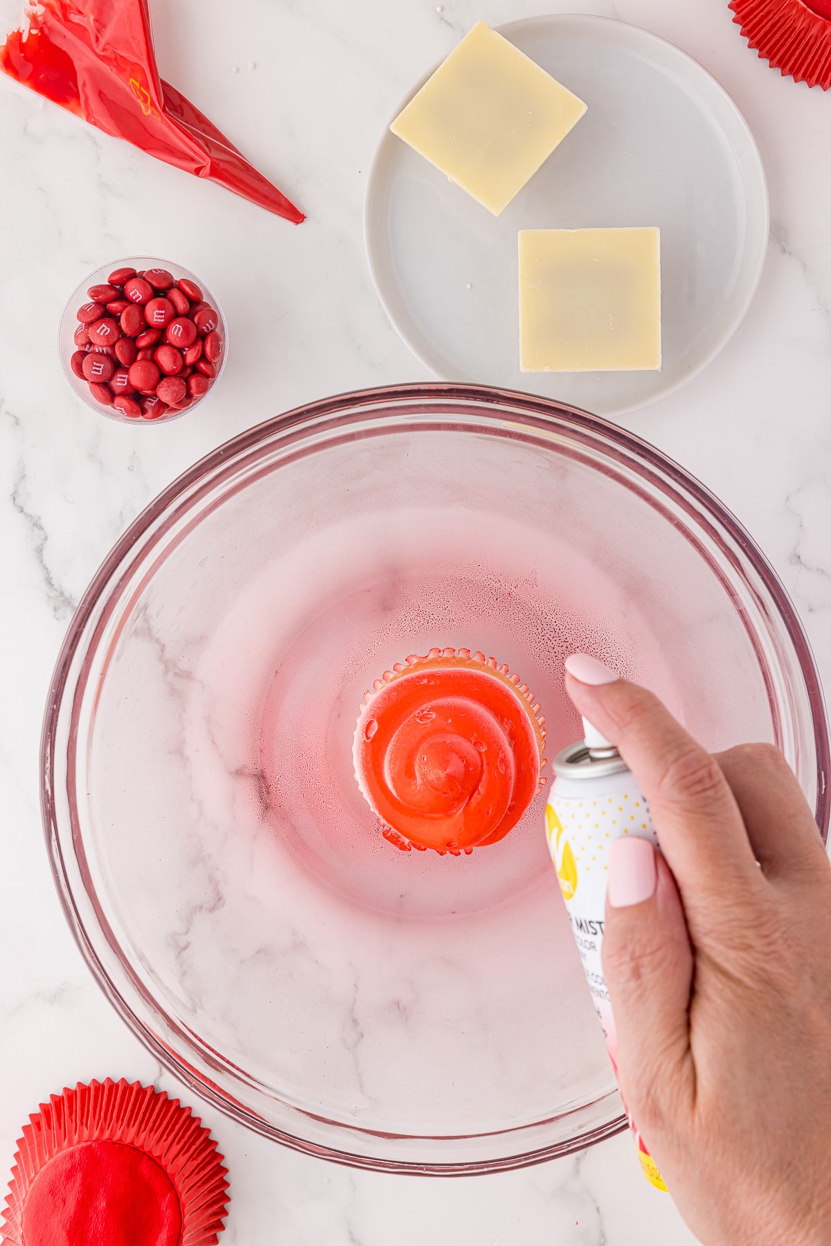 A cupcake in a bowl being sprayed with Wilton red food coloring spray