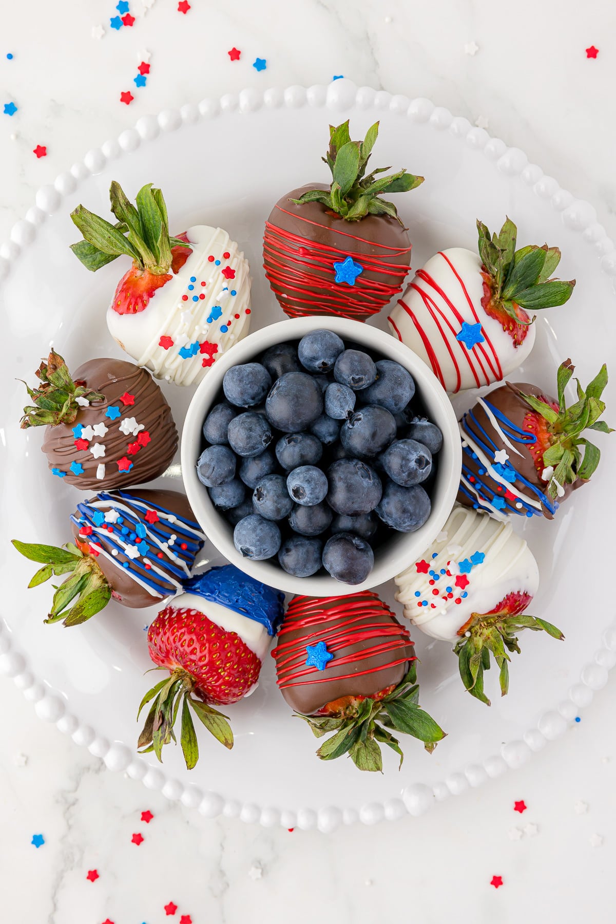 9 chocolate covered strawberries decorated in red, white and blue, displayed on a round white plate with a small white bowl of blueberries in the middle of the plate and the strawberries surrounding the bowl