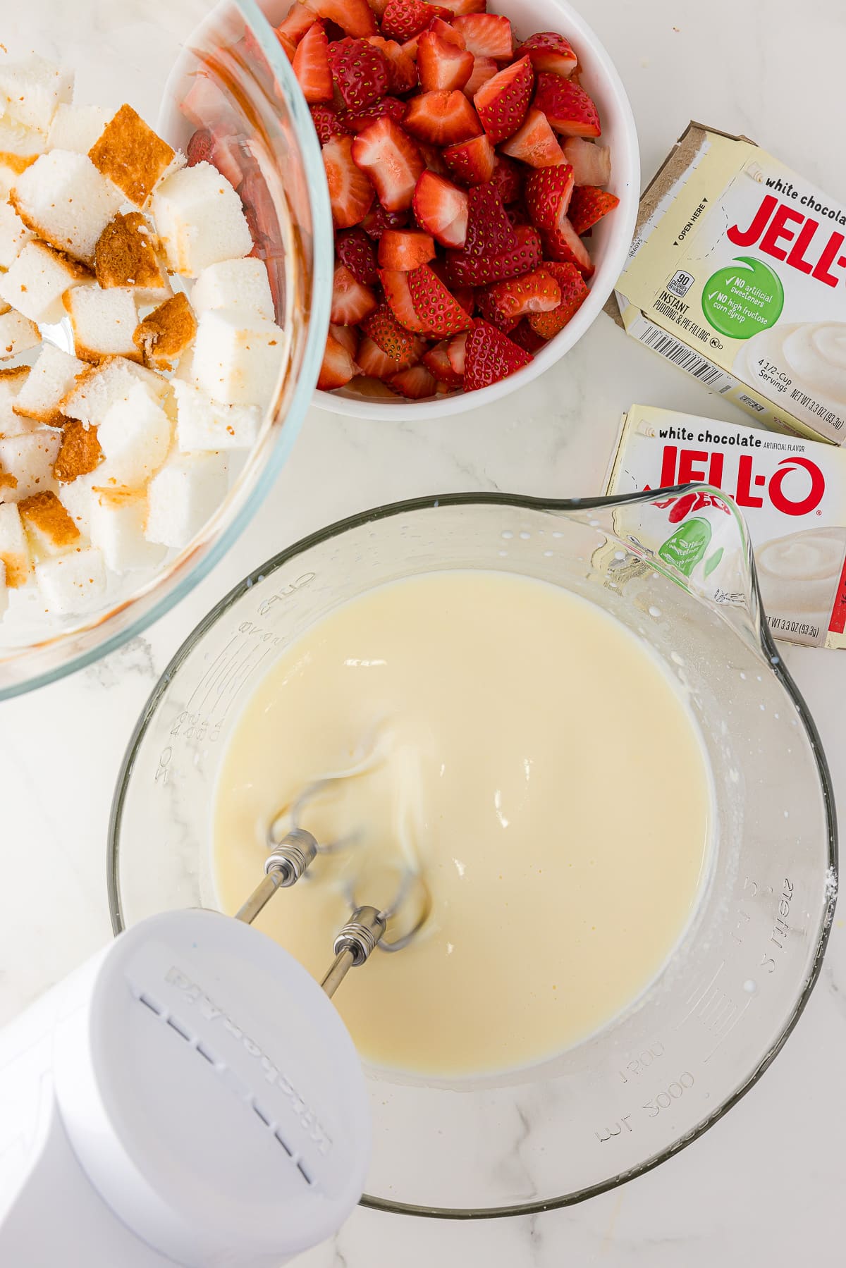 Hand held mixer in a large measuring cup mixing white chocolate pudding, with white chocolate pudding boxes, a bowl of strawberries, and a trifle bowl with a layer of angel food cake on the side