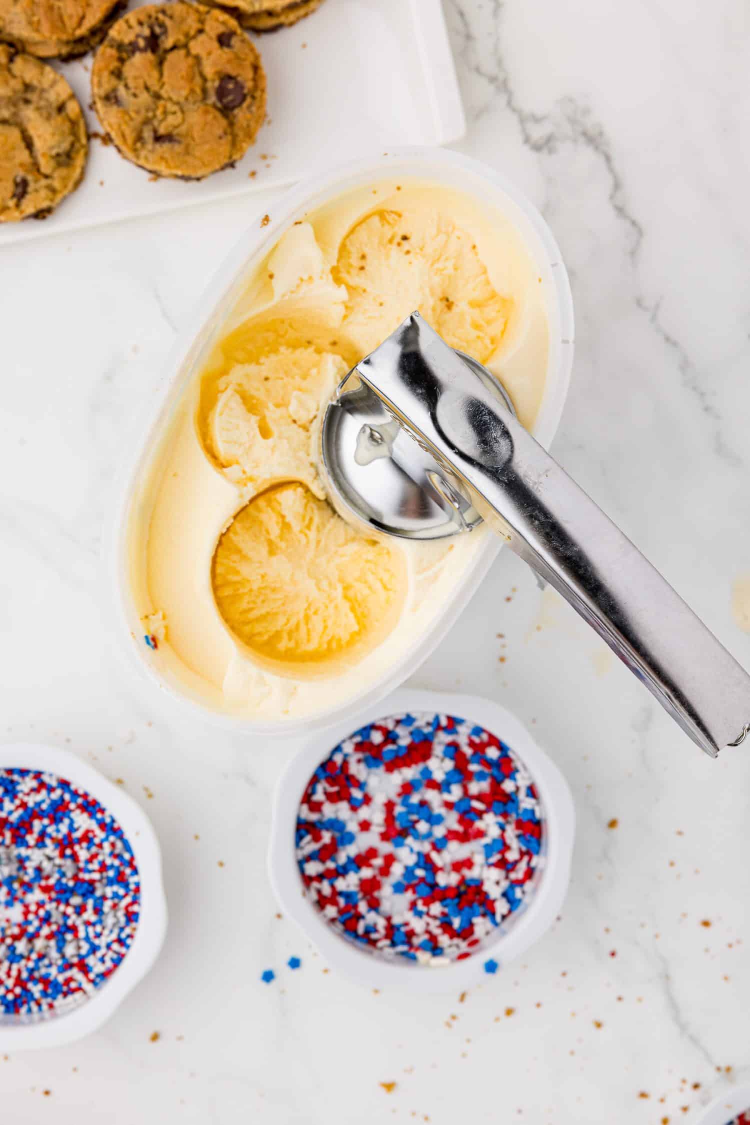 ice cream scoop in half gallon of ice cream with cookies and red white and blue sprinkles in the background