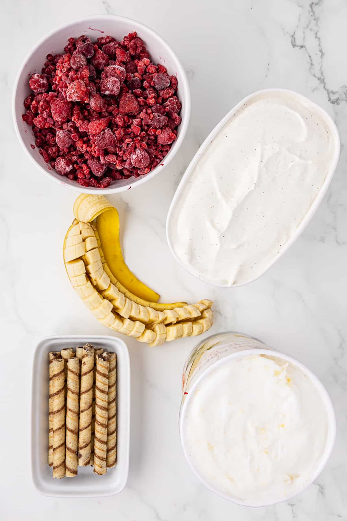 Ingredients for fresh fruit ice cream including container of vanilla ice cream, bowl of raspberries, whole banana peeled and cut into pieces, and tray of thin, tubular cookie wafers filled with chocolate
