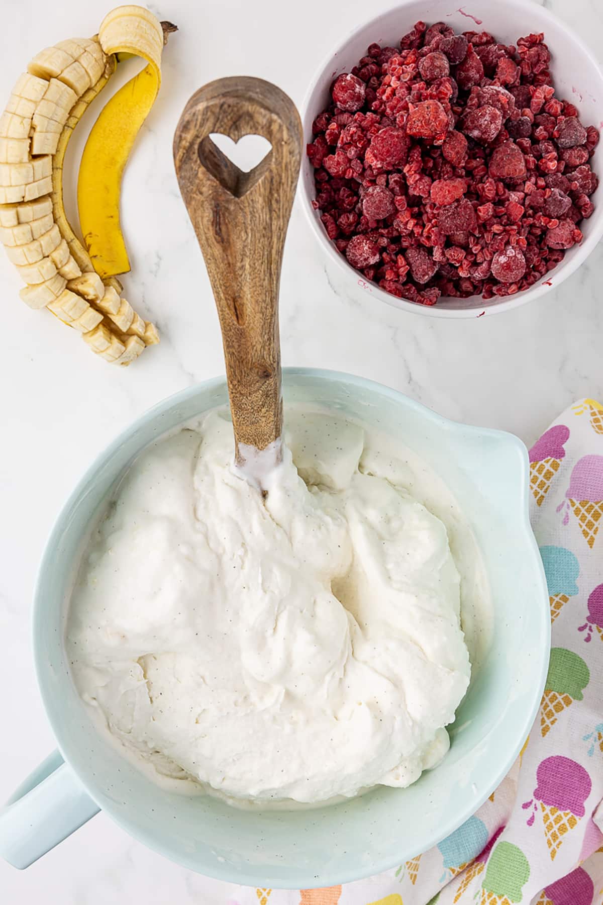 Large mixing bowl/cup with vanilla ice cream and wooden spoon in it, surrounded by whole banana cut in pieces, and a bowl of fresh raspberries