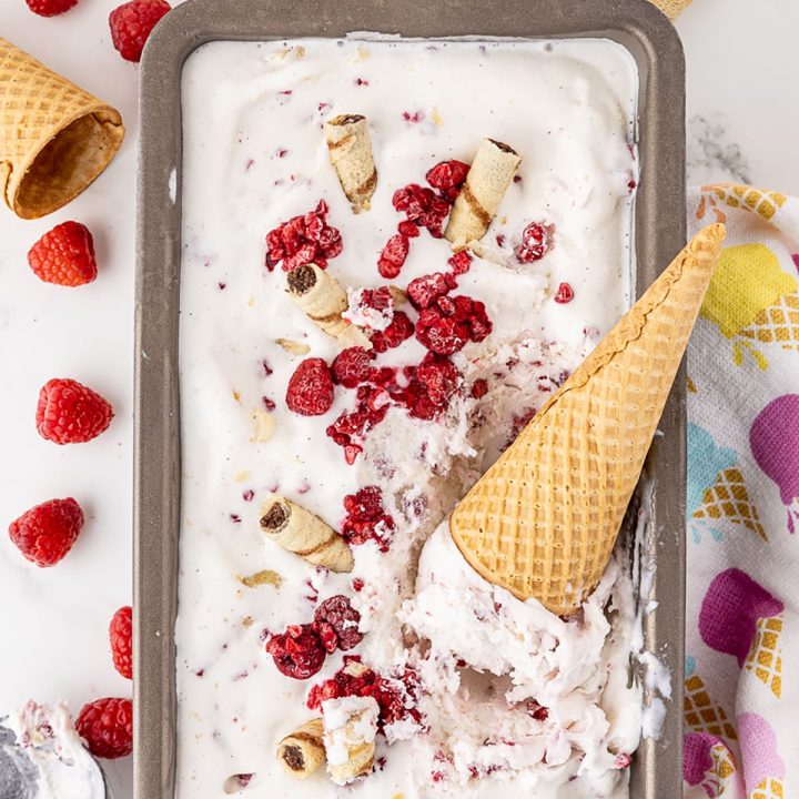 Rectangular, metal container of fruit filled ice cream with ice cream in a sugar cone on top of the container. Container surrounded by ice cream scoop, fresh banana and raspberries, and sugar cones and thin, tubular wafer cookies