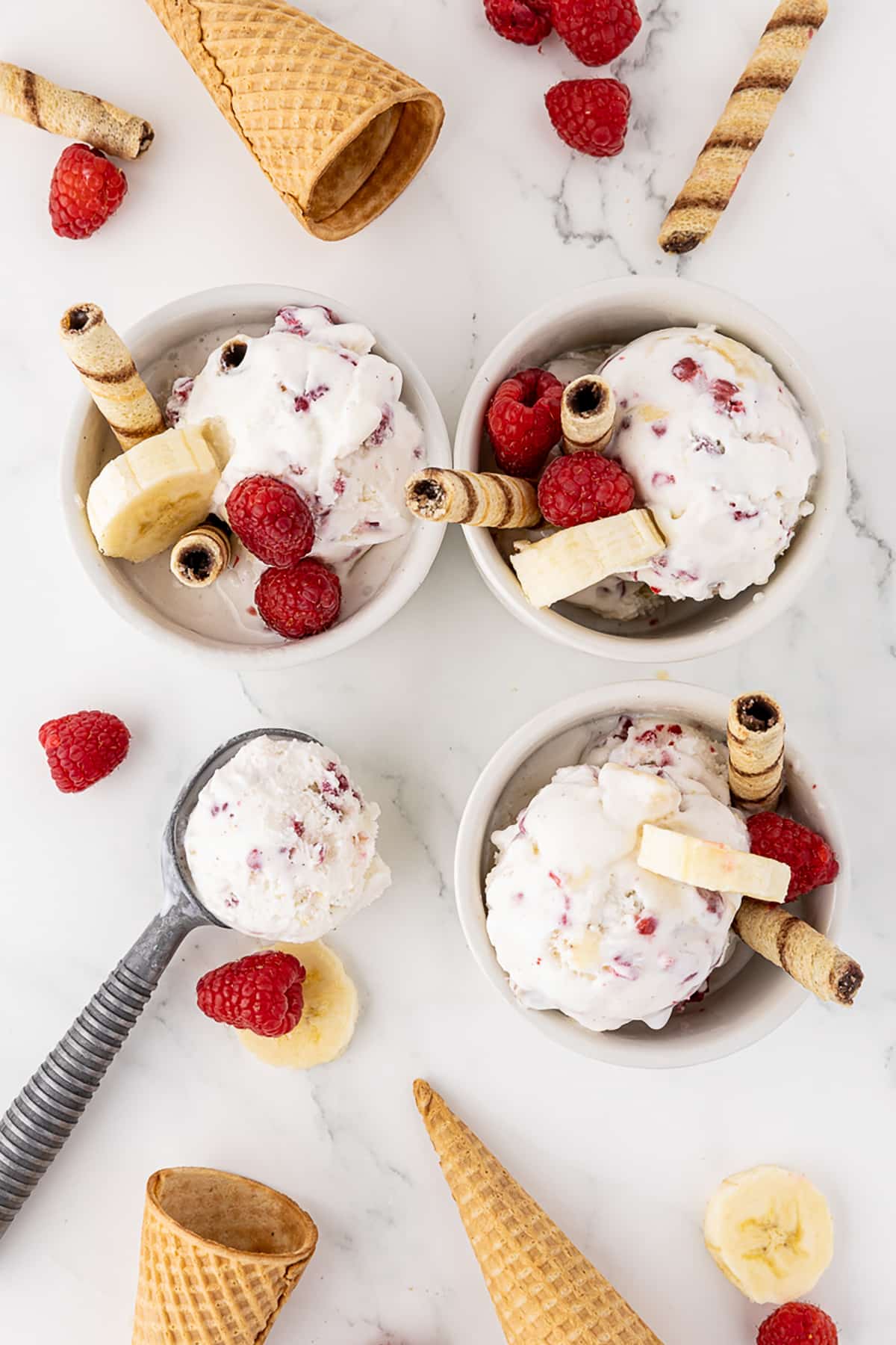 Three bowls of ice cream with raspberries and banana slices and tubular, spiraled cookies surrounded by ice cream cones, fresh bananas and strawberries and an ice cream scoop with a scoop of ice cream