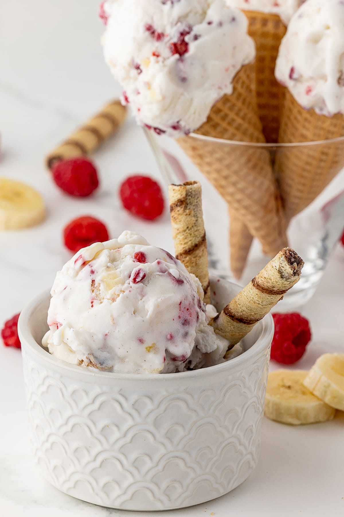 Bowl of ice cream with thin tubular cookie wafers, and three sugar cones filled with raspberry and banana fresh fruit ice cream, all standing in a large cone shaped glass in the background