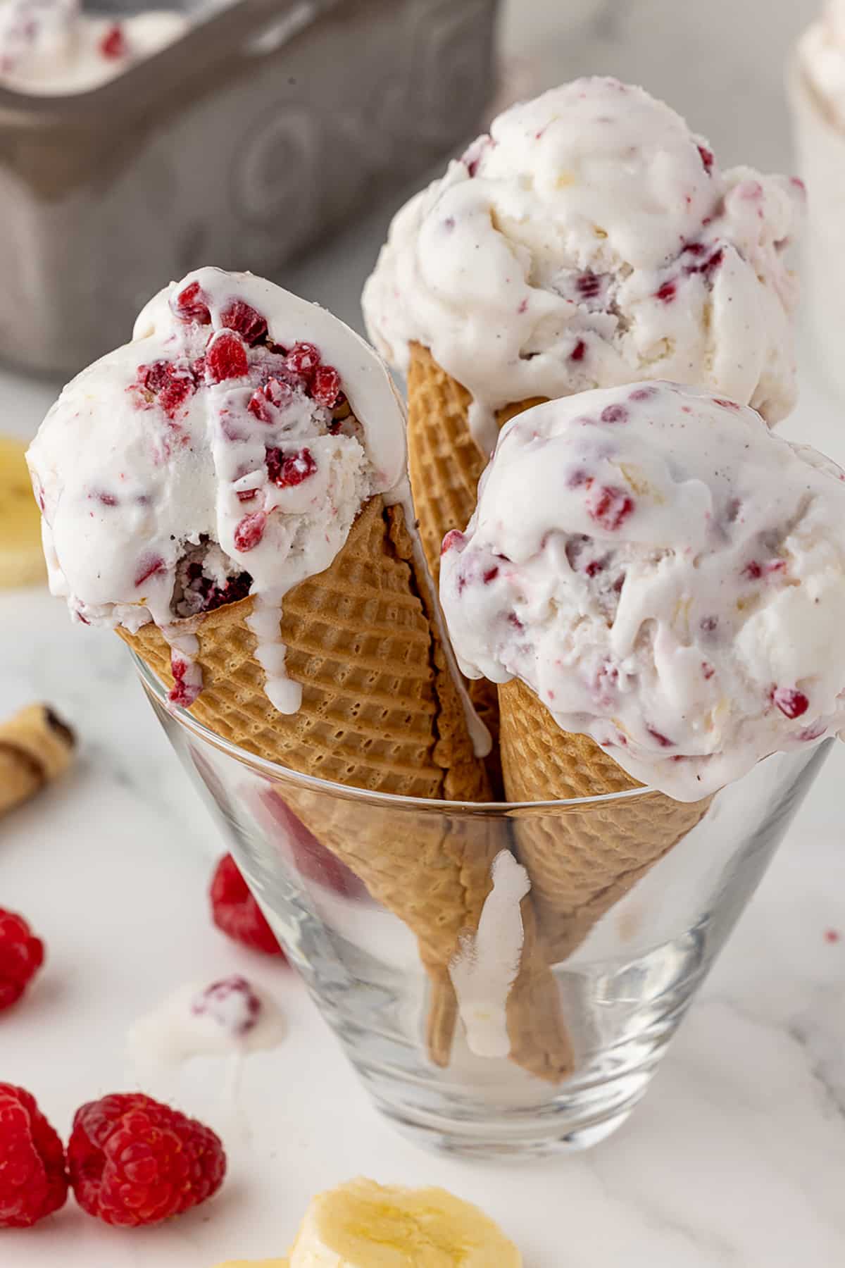 Close up of three sugar cones filled with raspberry and banana fresh fruit ice cream, all standing in a large cone shaped glass, with fresh raspberries and bananas in the foreground and a metal, rectangular container 