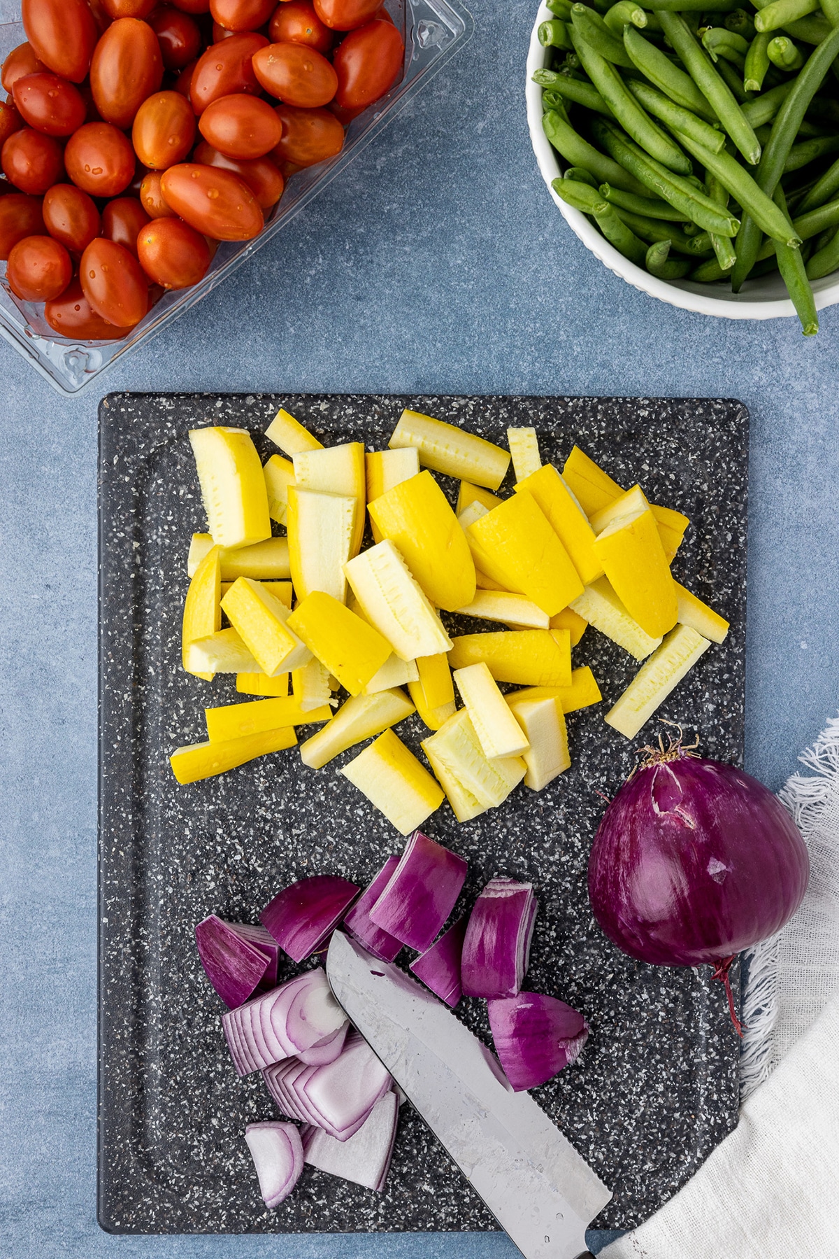 Chopped red onions and yellow squash on a cutting board, with a bowl of green beans and a container of cherry tomatoes in the background. 