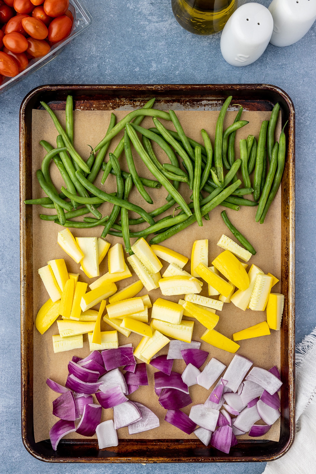 Sheet pan with green beans, yellow squash, and red onions on a blue countertop. 