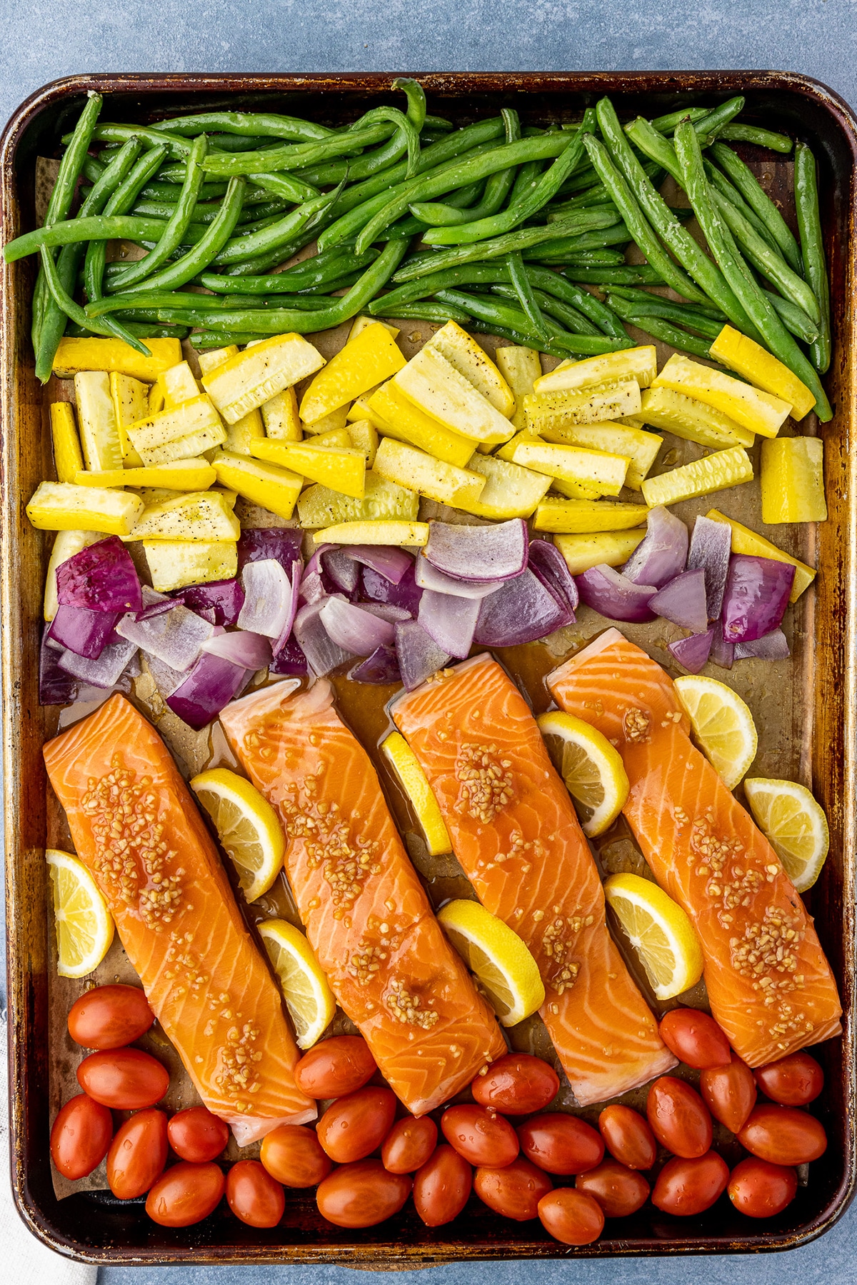 Sheet pan with a row of green beans, yellow squash, red onions, salmon filets, and cherry tomatoes, ready to be put in the oven.