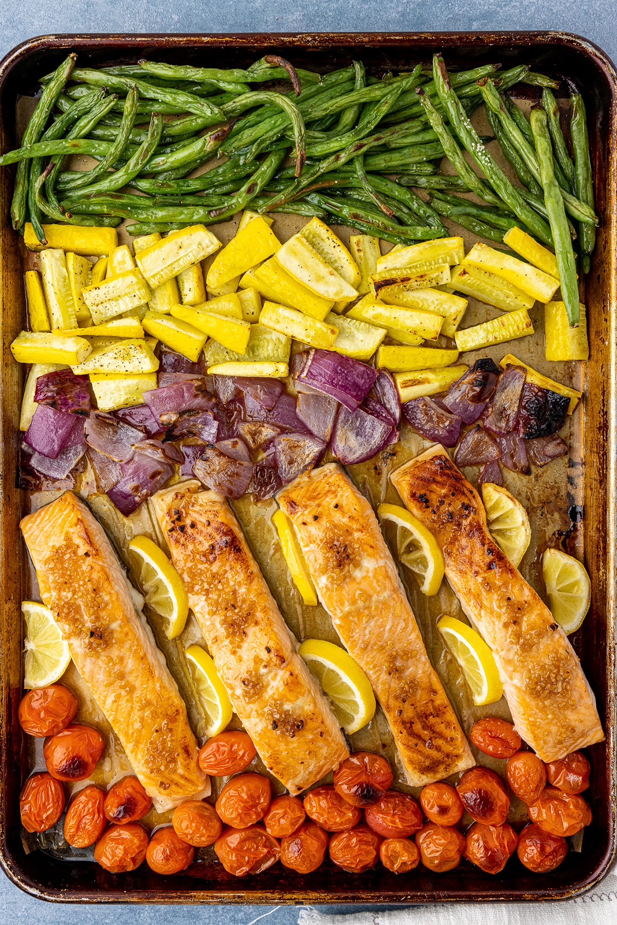 Sheet pan with a row of green beans, yellow squash, red onions, salmon filets, and cherry tomatoes.