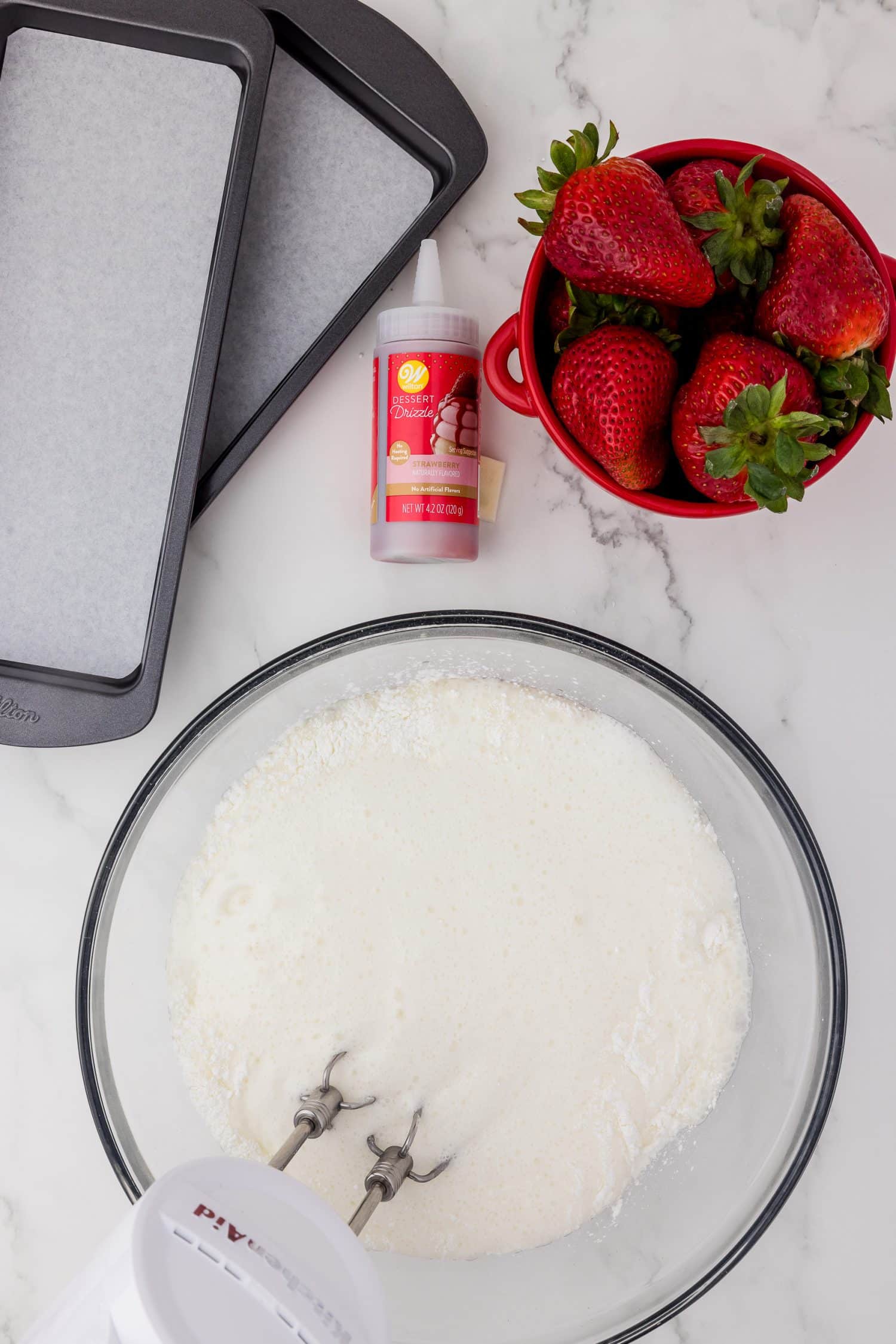 Mixing white batter in a glass blow with a red bowl filled with strawberries and loaf pans lined with parchment.