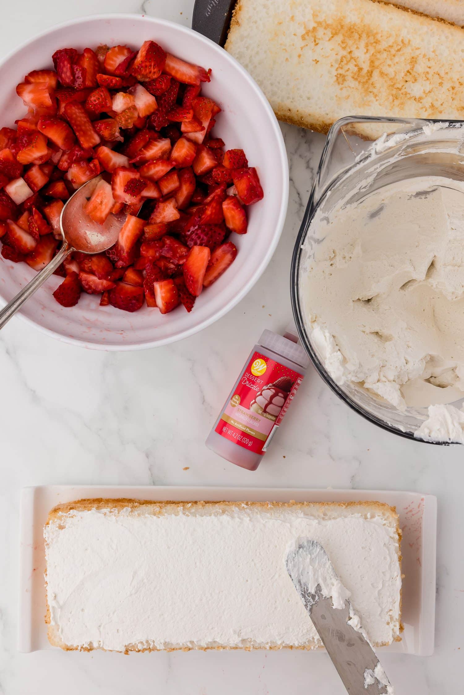 spreading whipped cream on a layer of angel food cake, bowl of strawberries, bowl of whipped cream and Wilton strawberry dessert drizzle on a white marble countertop