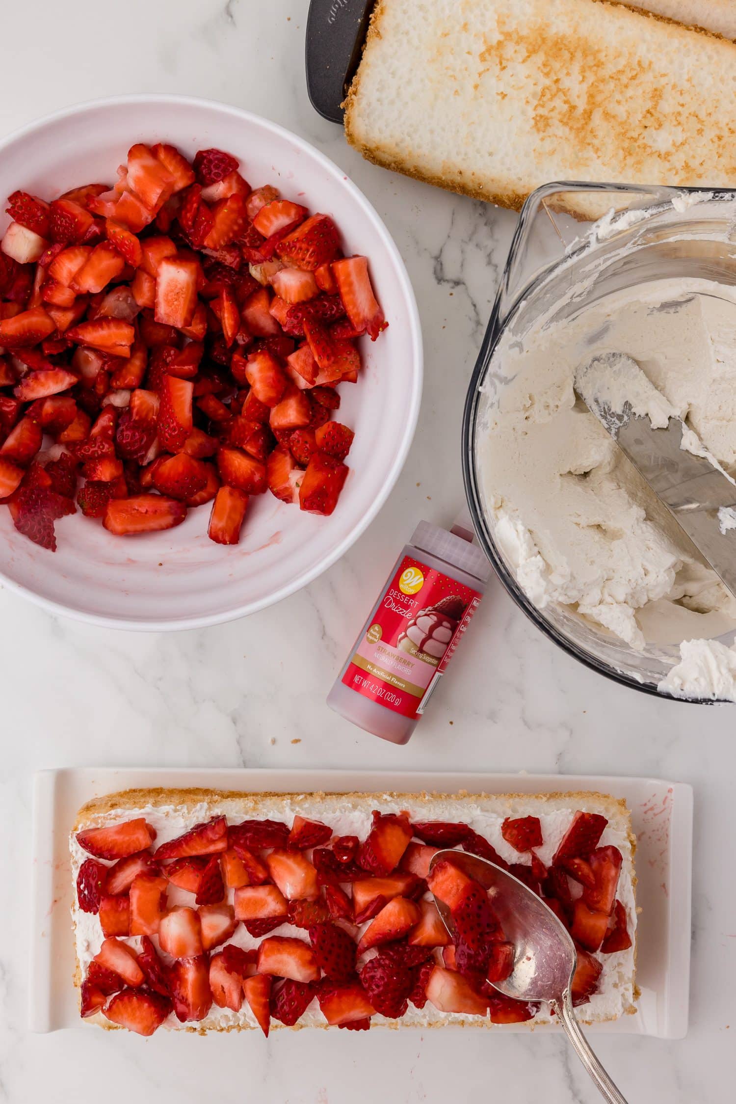 placing diced strawberries on top of a layer of angel food cake, a bowl of strawberries, whipped cream, and Wilton Dessert Drizzle on a white marble countertop.