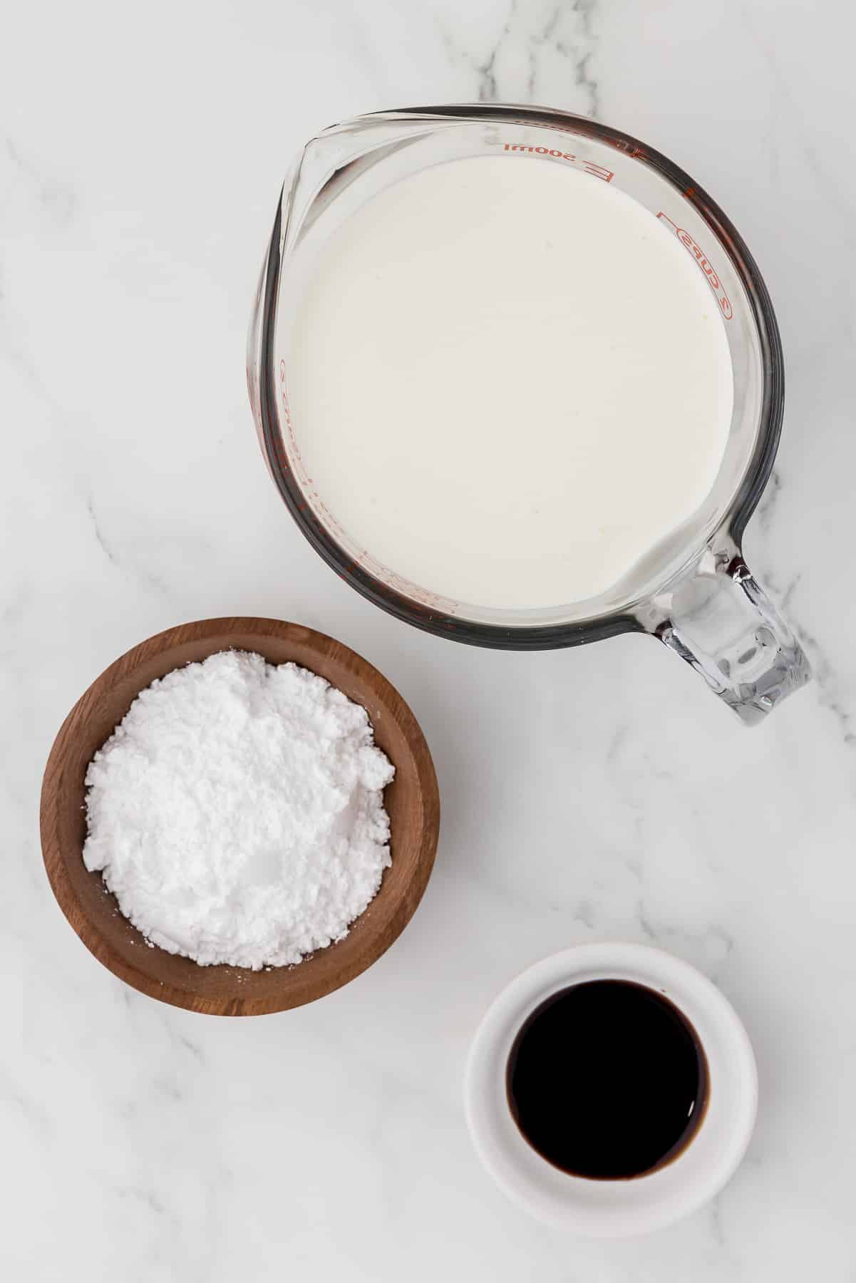 Mixing bowl of heavy cream, powdered sugar, and vanilla on a white countertop.