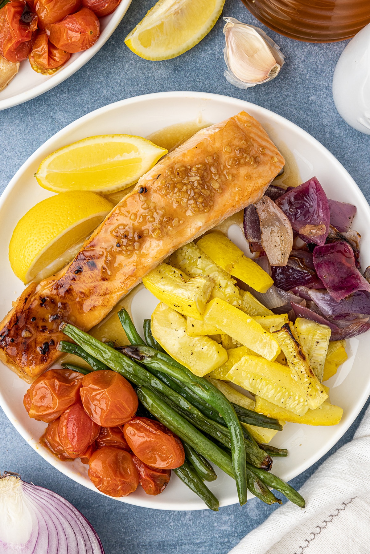 Green beans, yellow squash, red onions, salmon filets, and cherry tomatoes, served on a white, round plate. 