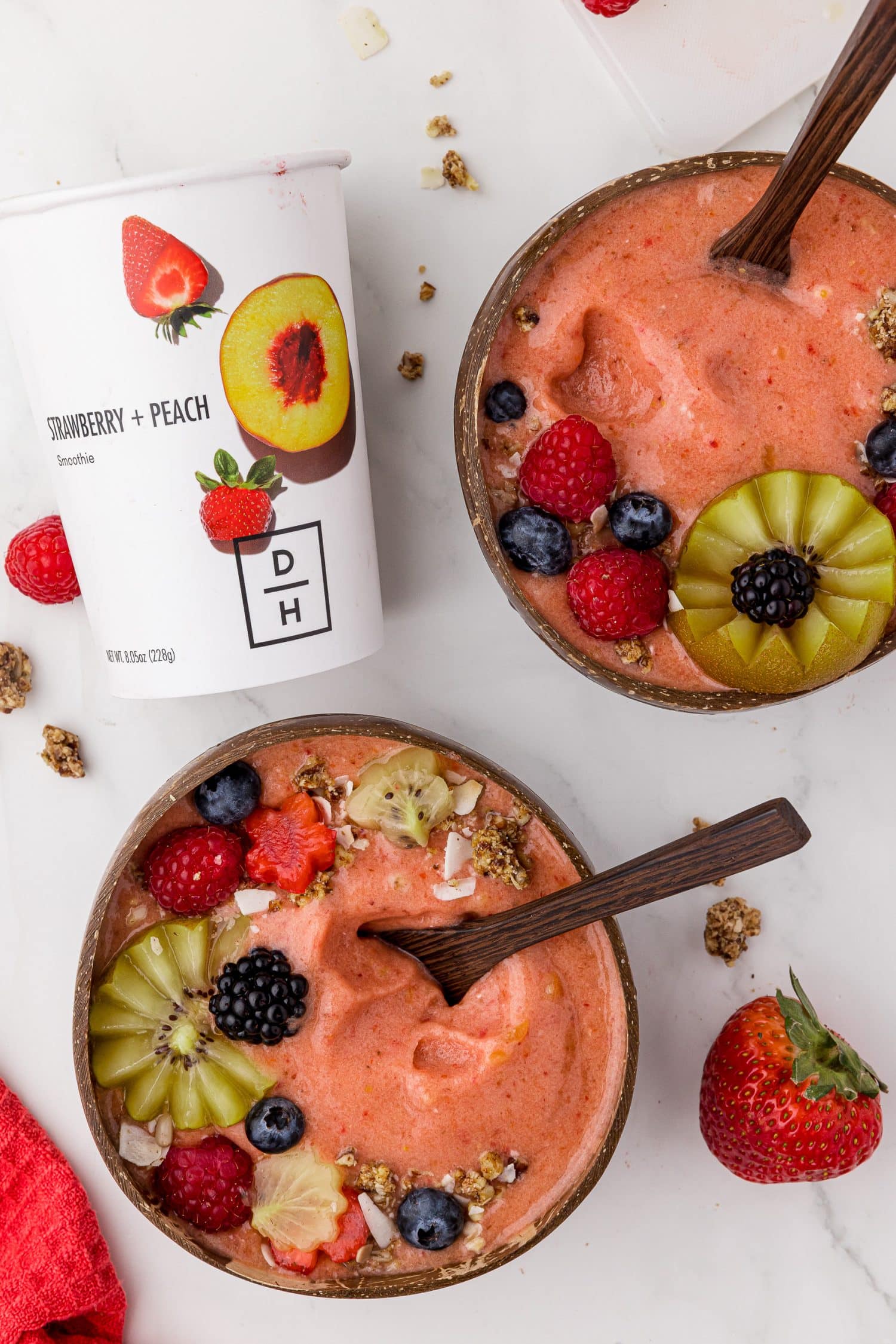 two coconut bowls filled with fruit smoothie and fresh berries on a white countertop, with a daily harvest cup
