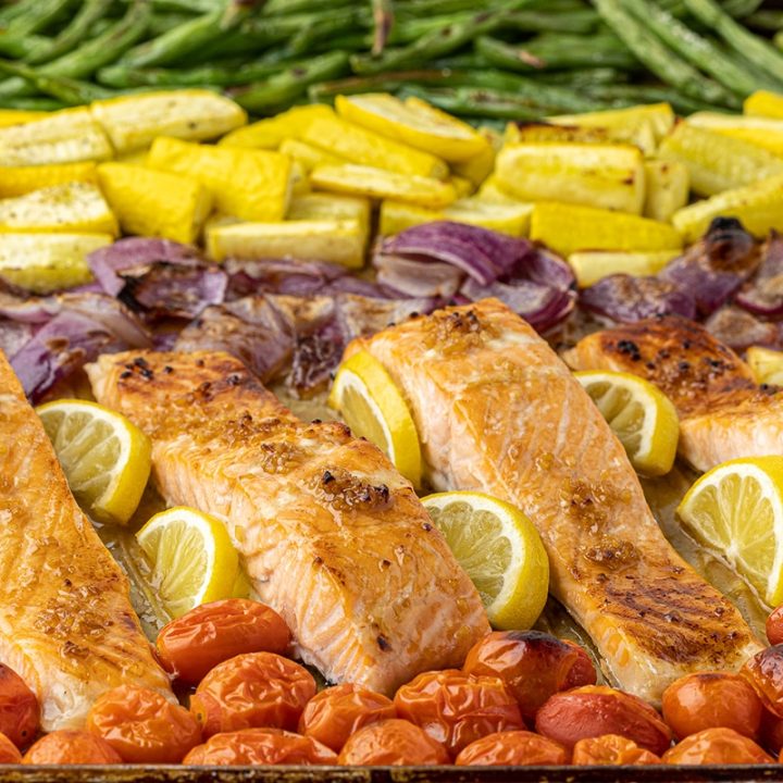 Cherry tomatoes, salmon filets, lemon wedges, red onions, yellow squash, and green beans on a sheet pan.
