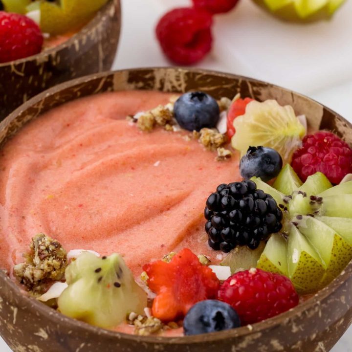 two coconut bowls filled with fruit smoothie and fresh berries on a white countertop with two raspberries and a kiwi in the background