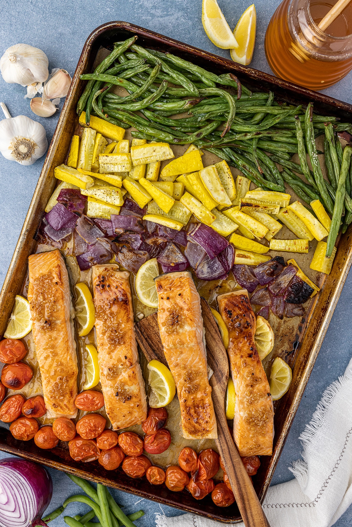 Sheet pan with a row of green beans, yellow squash, red onions, salmon filets, and cherry tomatoes, with a wooden serving spoon on a blue countertop. 