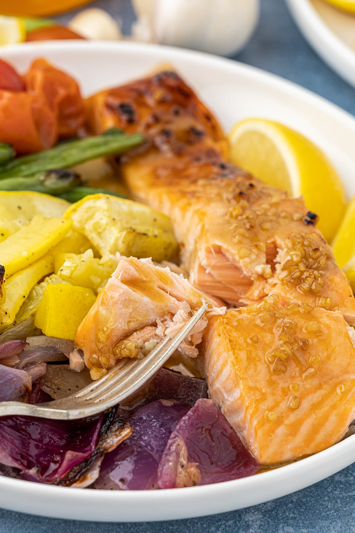 Closeup of a piece of salmon filet on a fork, with red onions and yellow squash on a round plate. 