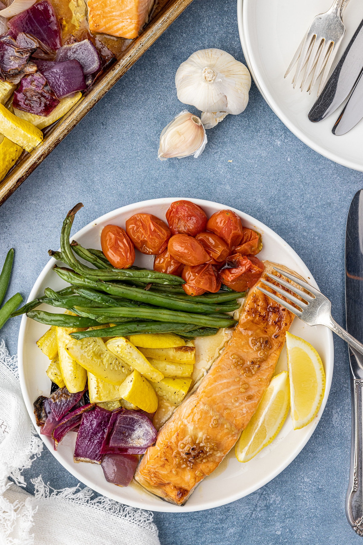 Salmon filet, green beans, cherry tomatoes, yellow squash, red onions, and lemon slices on a white, round plate on a blue countertop. 