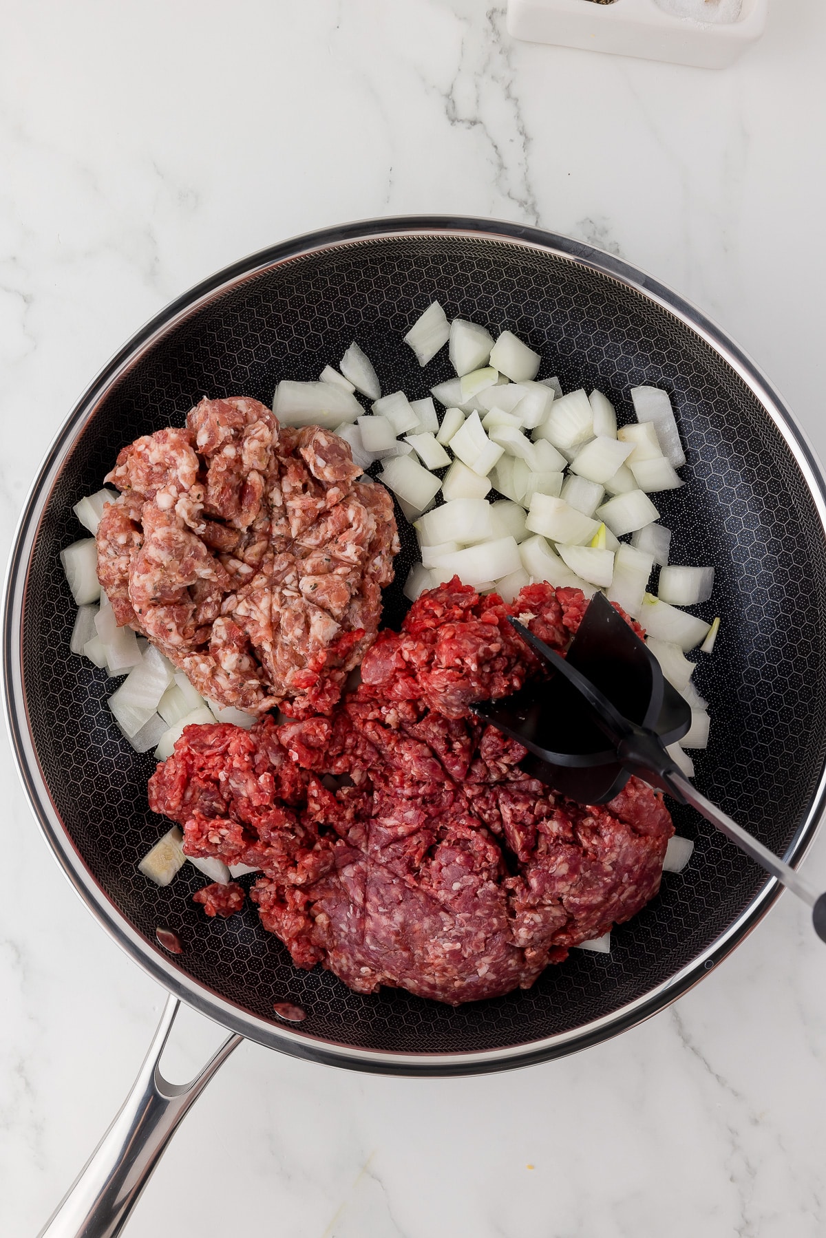 Raw ground meat, ground sausage, chopped onions in a Gordon Ramsay hexclad pan on a white marble countertop.